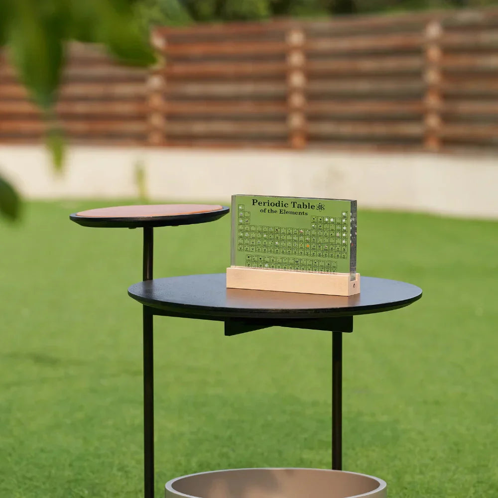 Periodic table on a small round table outdoors with green grass and wooden benches in the background
