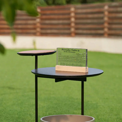 Periodic table on a small round table outdoors with green grass and wooden benches in the background