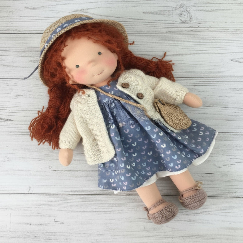 Doll with red hair wearing a blue dress with white patterns, beige sweater, and straw hat on a wooden surface.