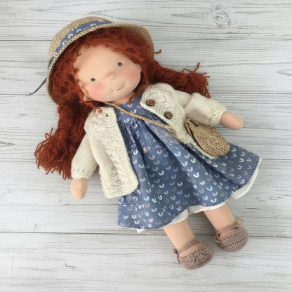Doll with red hair wearing a blue dress with white patterns, beige sweater, and straw hat on a wooden surface.