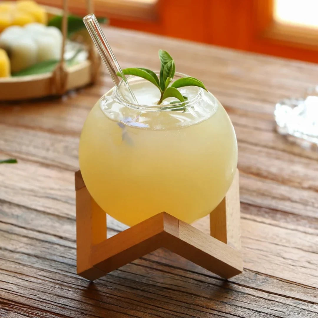 Josh & Tate - Floating Sphere Glass (16oz / 500mL) - Yellow drink in a glass with a straw and mint leaf on a wooden stand, set on a wooden table.