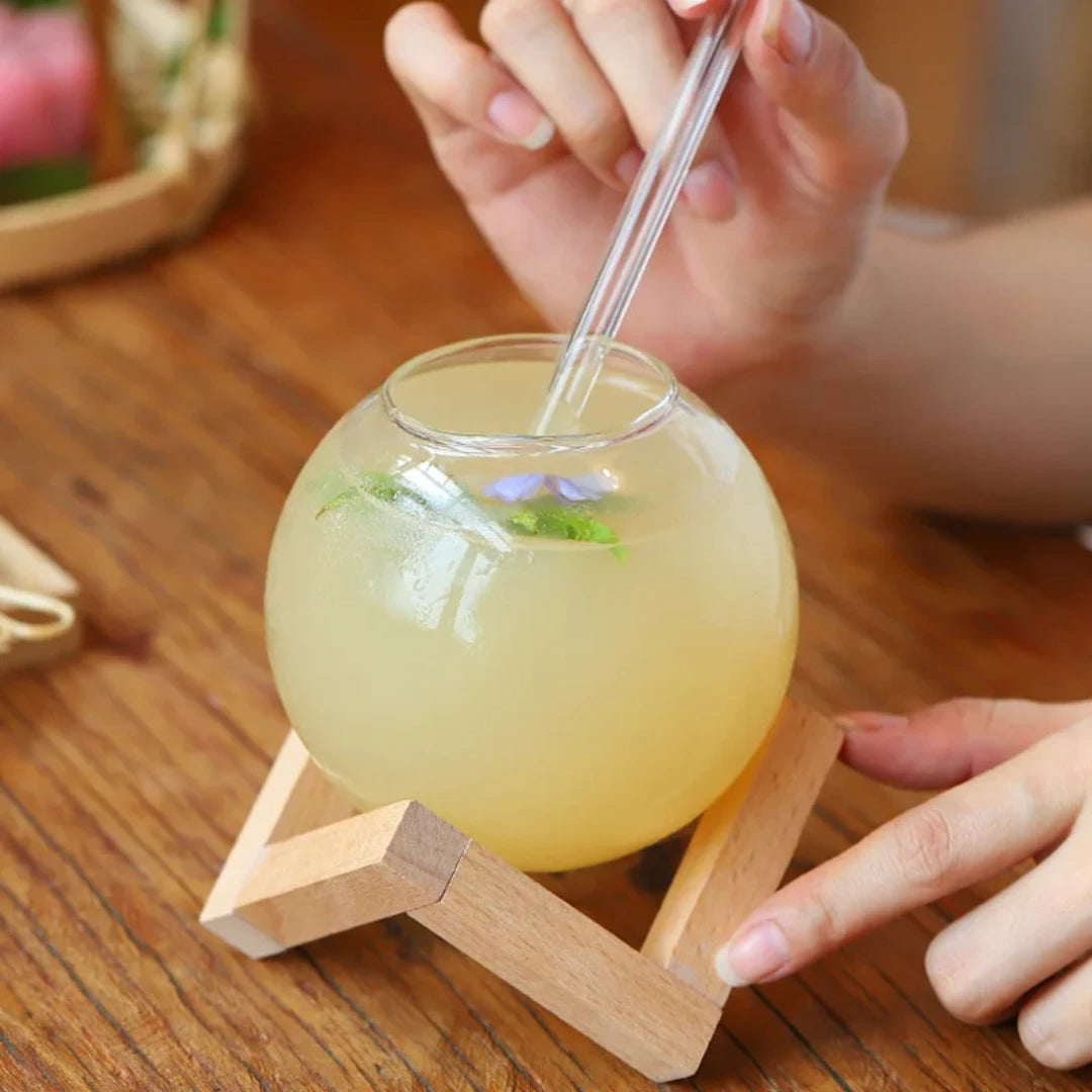 Josh & Tate - Floating Sphere Glass (16oz / 500mL) - Glass ball with yellow liquid and a straw on a wooden stand, held by a person.