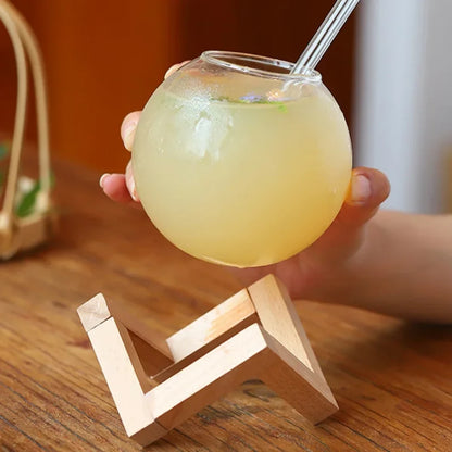 Josh & Tate - Floating Sphere Glass (16oz / 500mL) - Hand holding a glass of yellow liquid with a straw on a wooden stand.