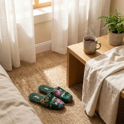 Josh & Tate - FloraStep™ Satin Fur-Lined Slipper - Cozy bedroom with floral slippers, a wooden side table with a mug, and a plant.