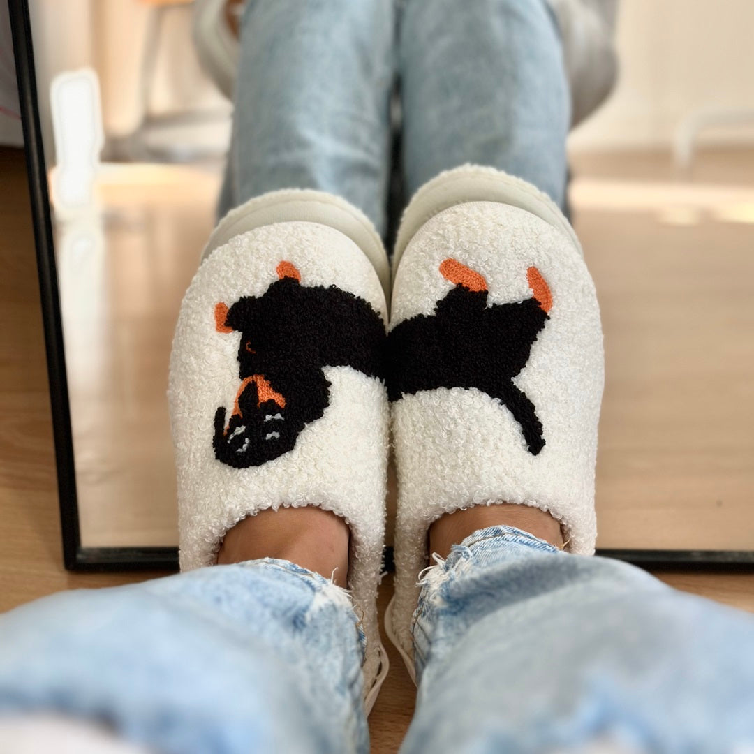 White slippers with black cat design worn by a person sitting on a couch.