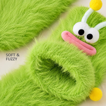 Josh & Tate - FuzzBuddy™ Cozy Socks - Green fuzzy monster slippers with large white eyes, a pink mouth, and yellow antennae, shown on a light neutral background with the text 'SOFT & FUZZY'.