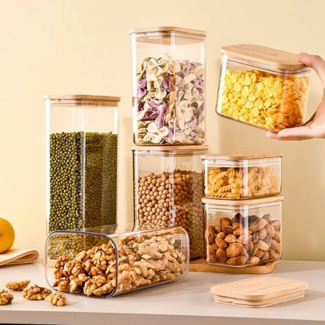 Set of clear storage jars with wooden lids containing various dry foods on a light surface.