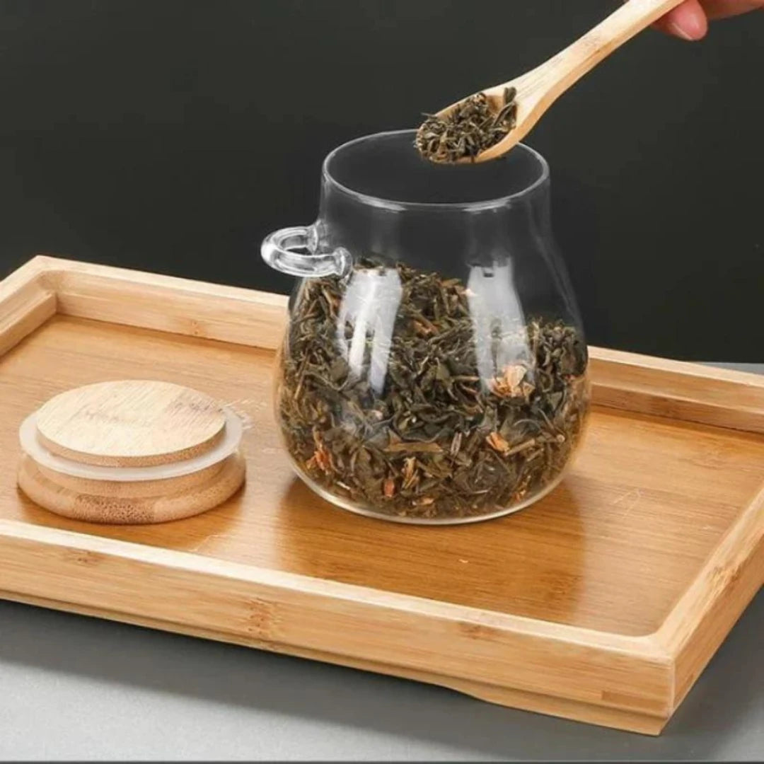 Josh & Tate - Glass Storage Jar with Spoon - Glass teapot with dry tea leaves on a wooden tray, with a hand holding a wooden spoon.