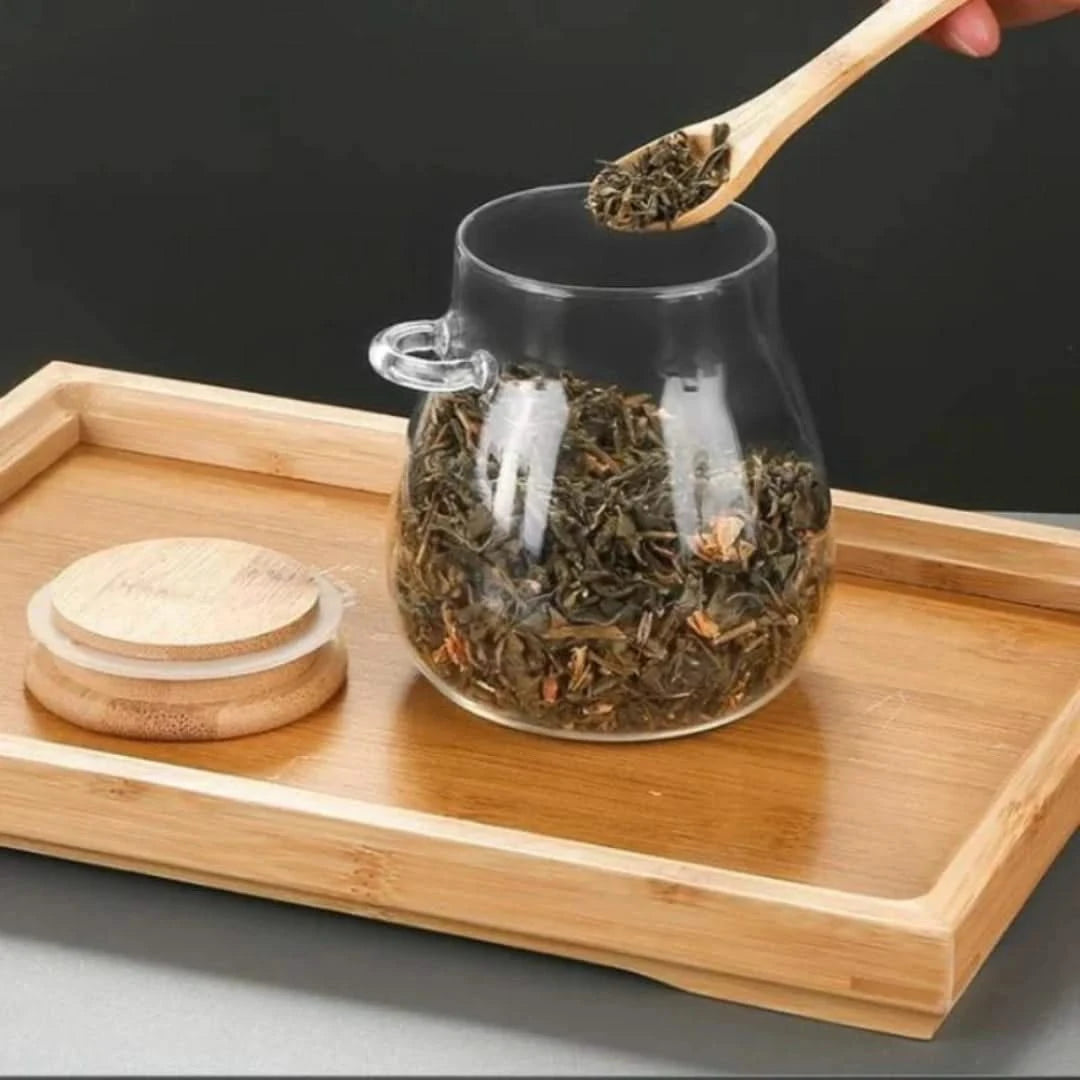 Josh & Tate - Glass Storage Jar with Spoon - Glass teapot with dry tea leaves on a wooden tray, with a hand holding a wooden spoon.