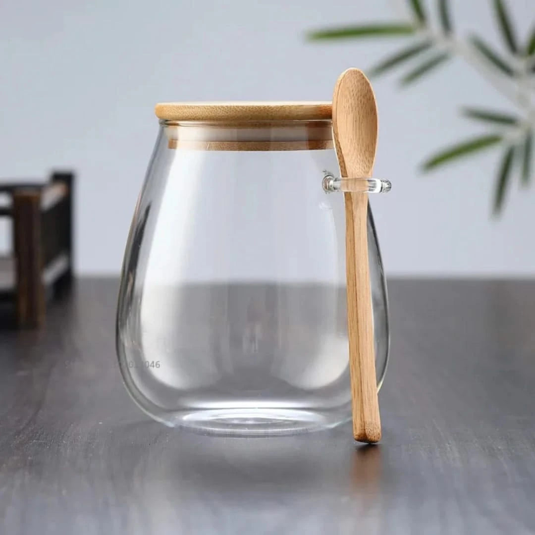 Josh & Tate - Glass Storage Jar with Spoon - Clear glass jar with wooden lid and spoon on a gray surface