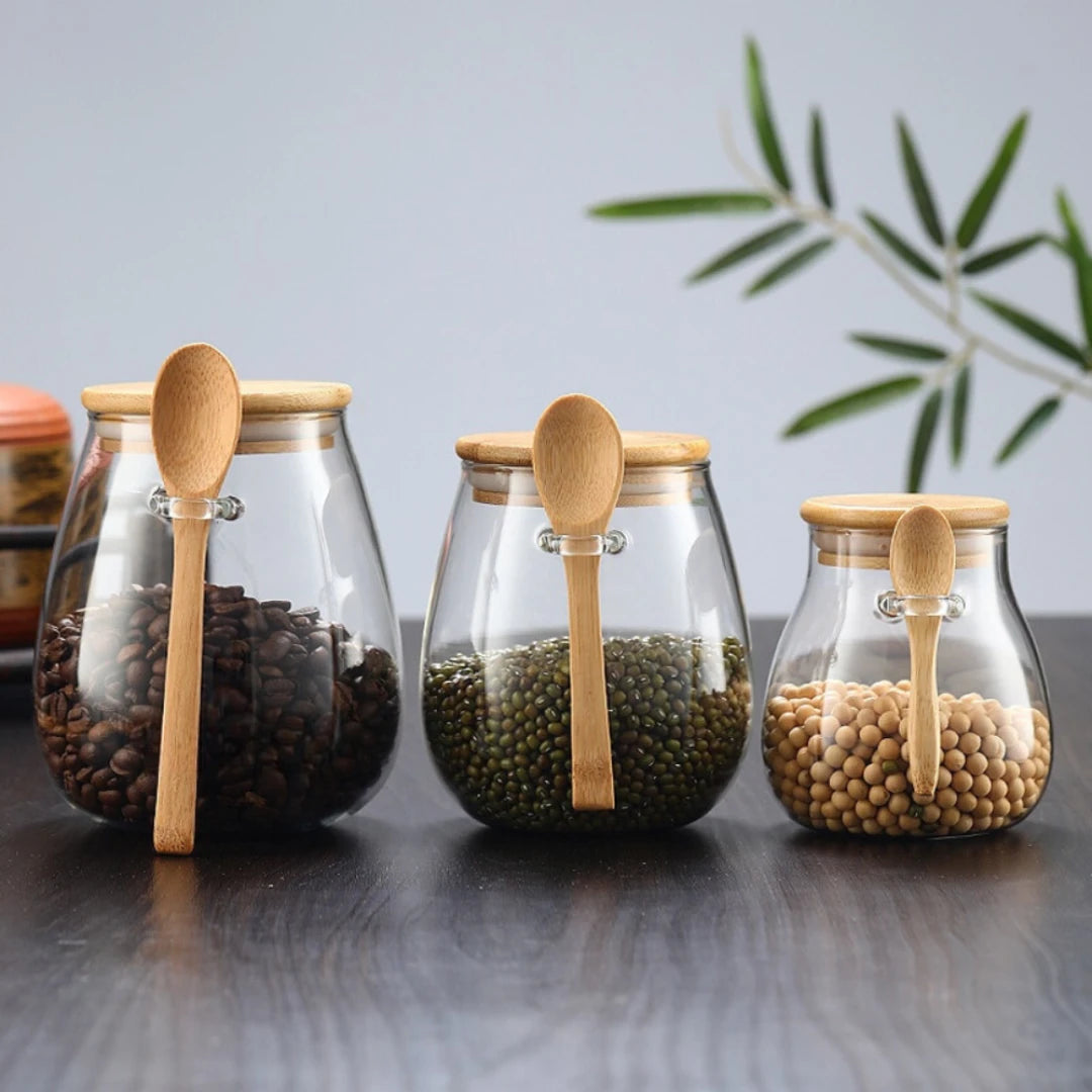 Josh & Tate - Glass Storage Jar with Spoon - Three glass jars with wooden spoons and lids, containing different types of beans on a reflective surface.