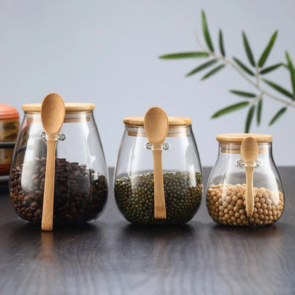 Josh & Tate - Glass Storage Jar with Spoon - Three glass jars with wooden spoons and lids, containing different types of beans on a reflective surface.