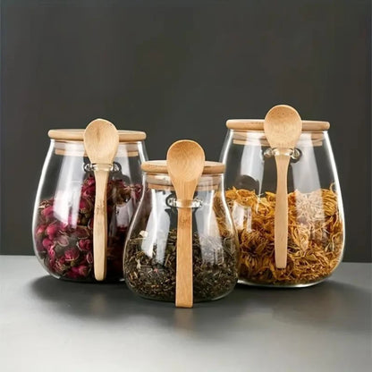 Josh & Tate - Glass Storage Jar with Spoon - Four glass jars with wooden spoons containing different types of tea leaves on a gray surface.
