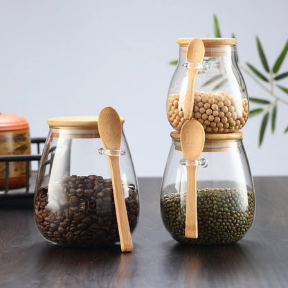 Josh & Tate - Glass Storage Jar with Spoon - Three glass jars with wooden lids and spoons containing different types of beans on a reflective surface.