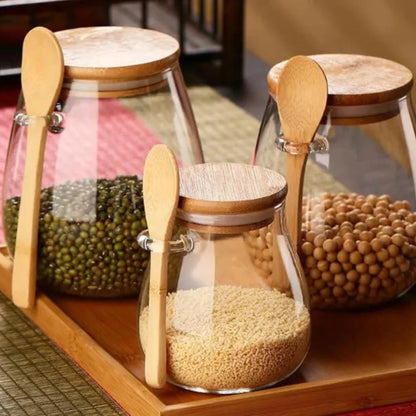 Josh & Tate - Glass Storage Jar with Spoon - Three glass jars with wooden lids and spoons on a wooden shelf, containing different types of grains.