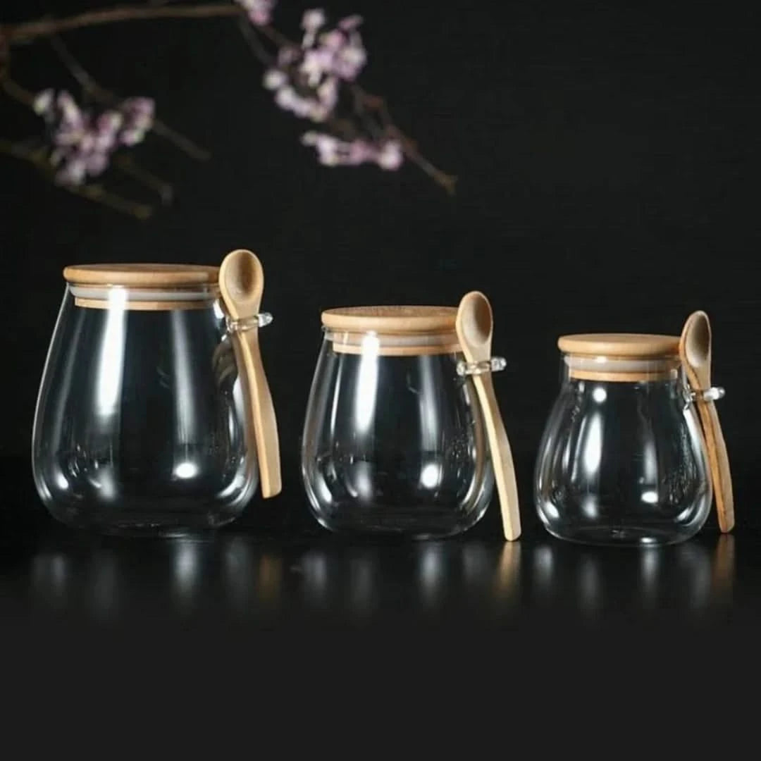 Josh & Tate - Glass Storage Jar with Spoon - Three glass jars with wooden lids and spoons on a dark background