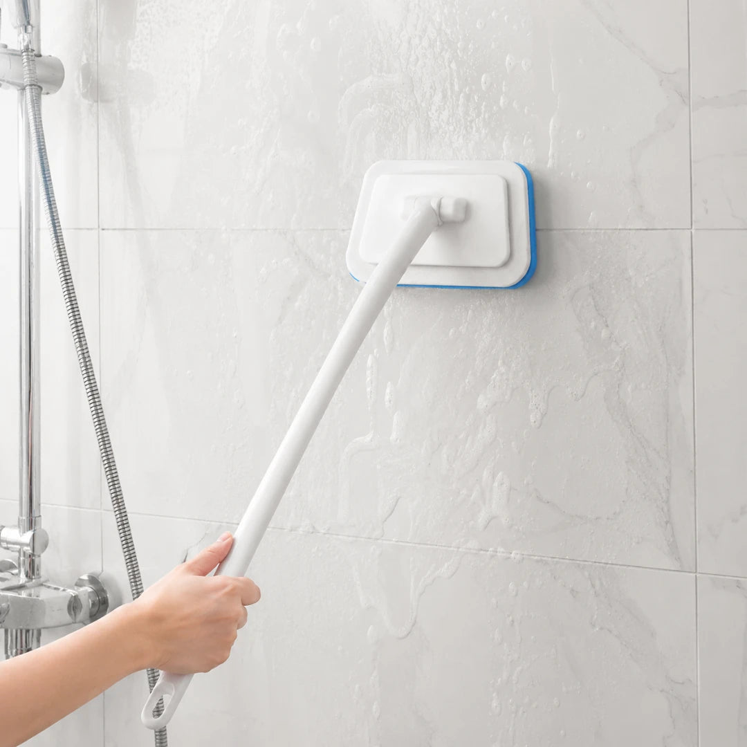 Josh & Tate - GlazeSafe™ Multi-Surface Scrub Brush - A hand holding a long white handle uses a wall-mounted cleaning brush with a blue edge to clean a tiled wall in a shower area.