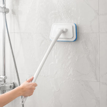 Josh & Tate - GlazeSafe™ Multi-Surface Scrub Brush - A hand holding a long white handle uses a wall-mounted cleaning brush with a blue edge to clean a tiled wall in a shower area.