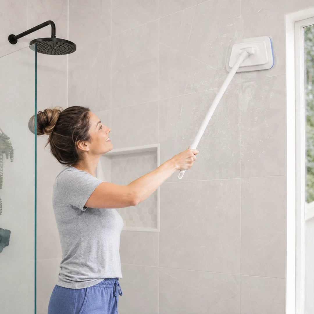 Josh & Tate - GlazeSafe™ Multi-Surface Scrub Brush - A woman in a gray t-shirt and blue pants uses a long-handled white shower wall cleaner with a blue-edged scrub pad to clean a tiled bathroom wall.