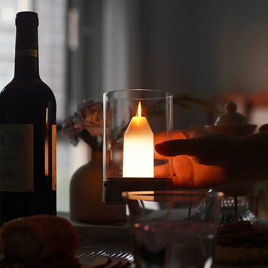 Candle in a glass holder on a table with a bottle of wine and a pumpkin in the background.