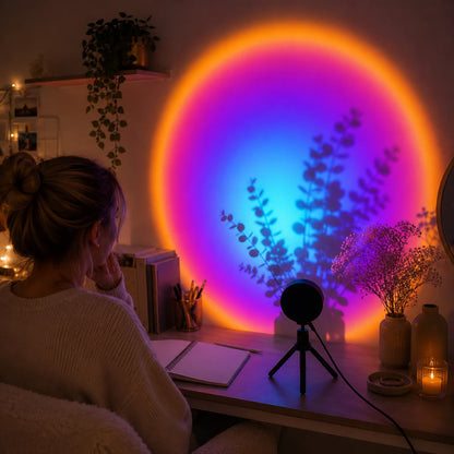 Josh & Tate - GoldenHour Sunset Lamp - A person sits at a desk in a dimly lit room, facing a large colorful circular light projection on the wall. The light displays a gradient of orange, yellow, pink, and blue, casting tree-like shadows on the wall.