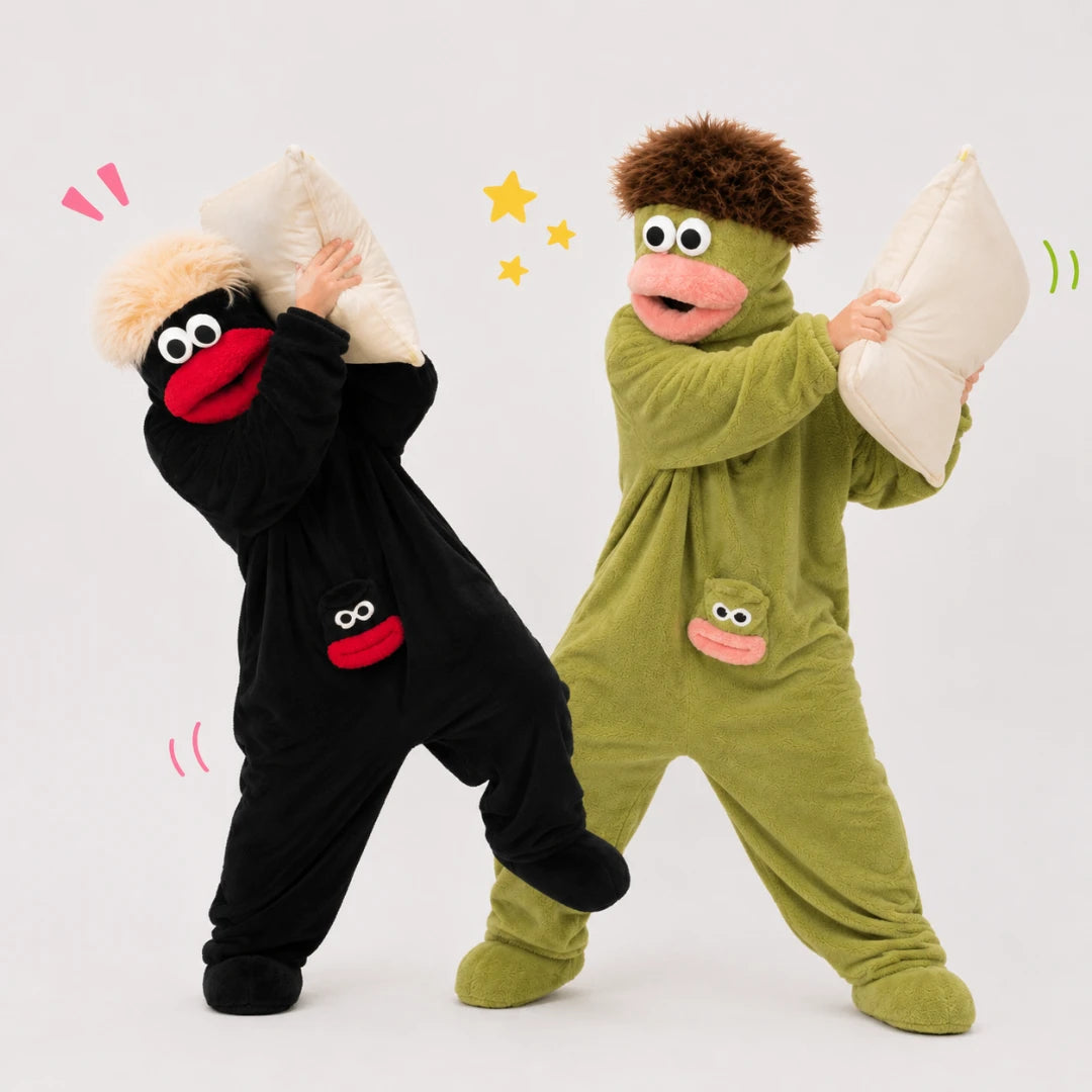 Josh & Tate - GoofSuit™ Big Mouth Monster Onesie - Two children wearing full-body costume pajamas pose playfully while holding white pillows against a light gray studio background. One costume is black with a red mouth and beige hair, and the other is olive green with a pink mouth and brown hair.