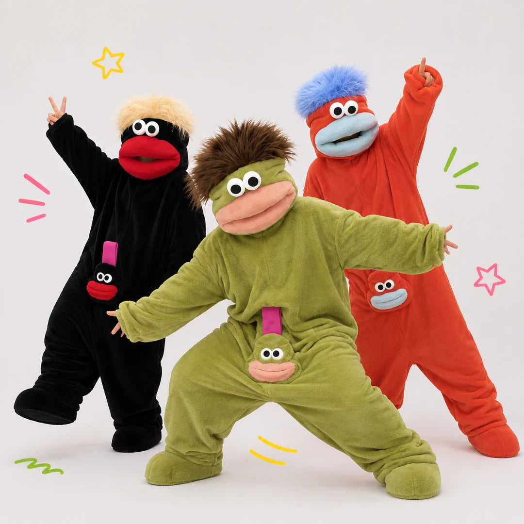 Josh & Tate - GoofSuit™ Big Mouth Monster Onesie - Three children wearing colorful monster costume suits posing together against a light gray studio background. The front child wears an olive green suit with brown hair and a pink mouth, the left child wears a black suit with a beige wig and red mouth, and the right child wears a red-orange suit with blue hair and a blue mouth.