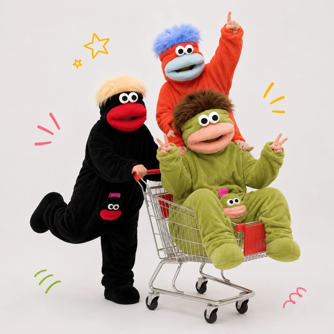 Josh & Tate - GoofSuit™ Big Mouth Monster Onesie - Three children wearing colorful monster and cartoon character costumes pose with a small shopping cart on a light gray studio background. One child is in a black costume with a red mouth, one in a green costume sitting in the cart, and one in an orange costume standing behind with a blue fluffy hat.