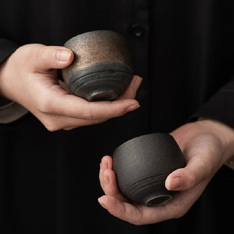 Two hands holding two dark ceramic cups against a black background