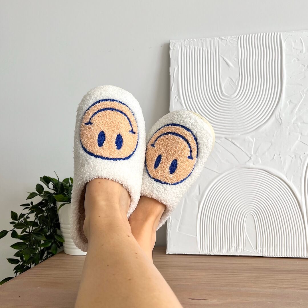 White slippers with smiley face design worn by a person, against a light gray wall with decorative patterns.