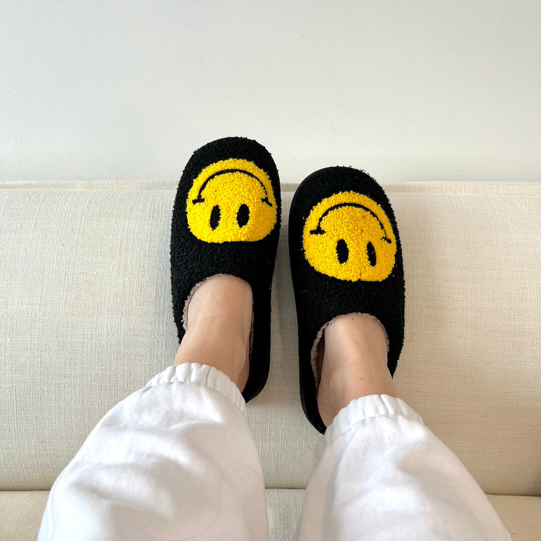 Black slippers with yellow smiley face design worn by a person on a light-colored couch.