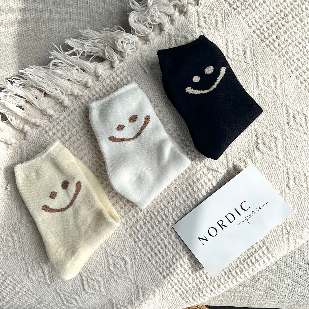 Three pairs of socks with smiley face designs on a textured surface, with a card labeled 'Nordic Place'.