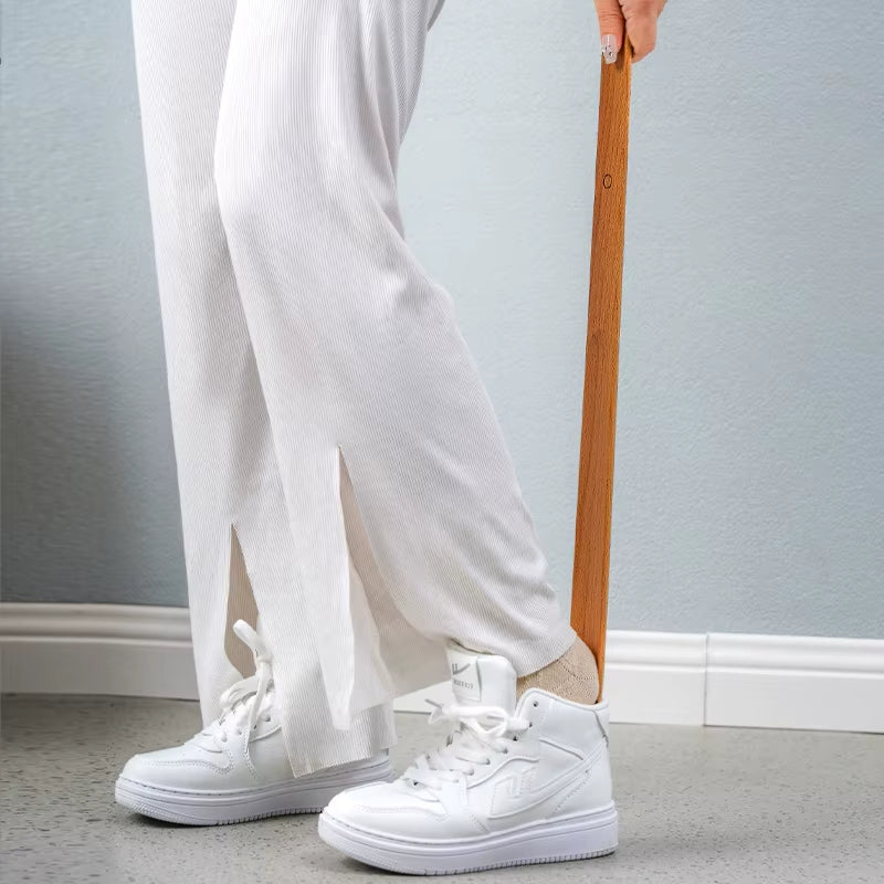 Person wearing white sneakers and light-colored pants holding a wooden object against a plain background