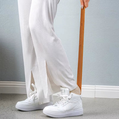 Person wearing white sneakers and light-colored pants holding a wooden object against a plain background