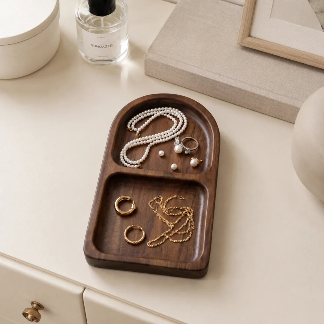 A dark brown wooden jewelry organizer tray with two compartments, styled on a cream-colored tabletop with various jewelry items placed inside.