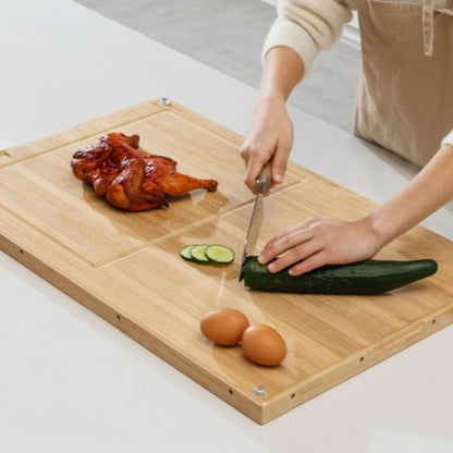 Person cutting a cucumber on a wooden cutting board with a roasted chicken and eggs.