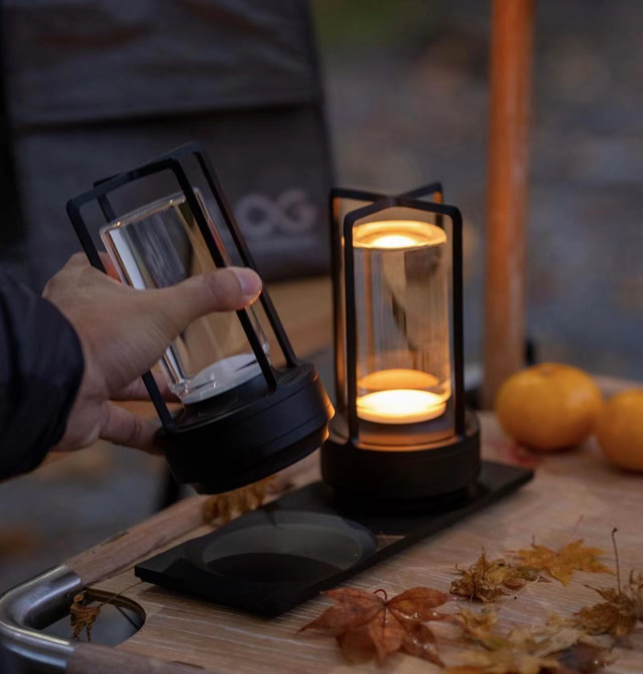 Person holding a lantern with another lantern on a table, surrounded by pumpkins and autumn leaves.