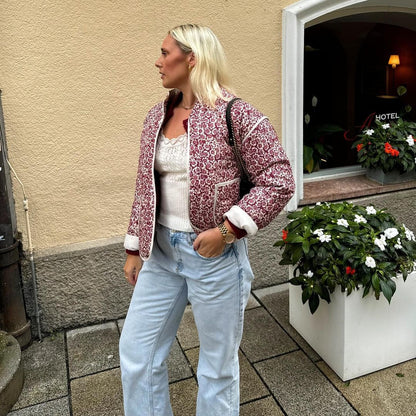 Woman in a red floral jacket and light blue jeans standing outside a building with flowers and a sign in the background.