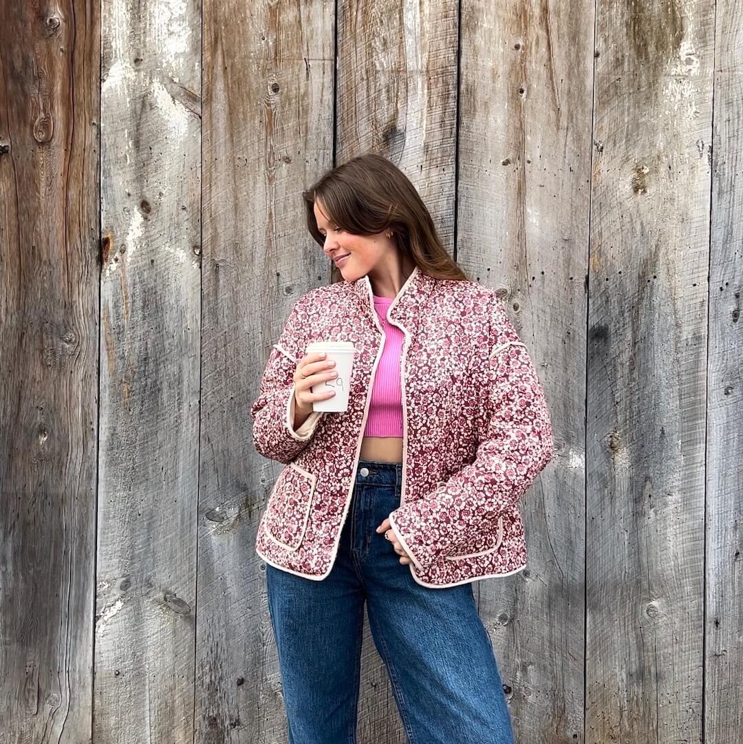 Woman wearing a pink floral jacket and blue jeans, holding a white mug against a wooden background