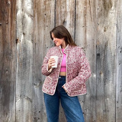 Woman wearing a pink floral jacket and blue jeans, holding a white mug against a wooden background