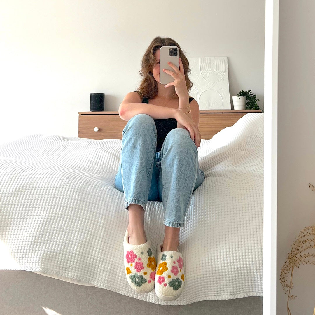 Person sitting on a bed wearing floral slippers, taking a mirror selfie.