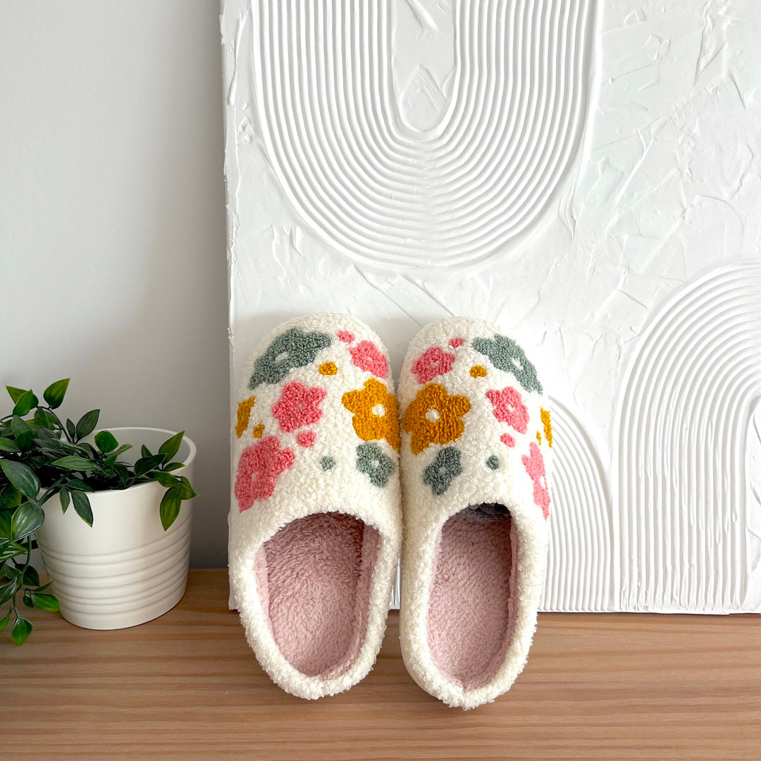 Pair of floral slippers on a wooden surface with a white textured wall in the background.