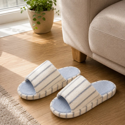 Josh & Tate - LoftWear™ Striped Slide Slippers - A pair of plush slide slippers with white and light blue stripes and a light blue footbed, placed on a wooden floor next to a beige sofa.