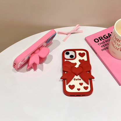 Josh & Tate - LoveBow™ Anti-Drop Phone Case - Two decorative phone cases displayed on a white tabletop: one pink case with a large pink bow and the other a red case with a red bow, both shown with matching pink ribbon accessories, alongside a pink book and a mug.