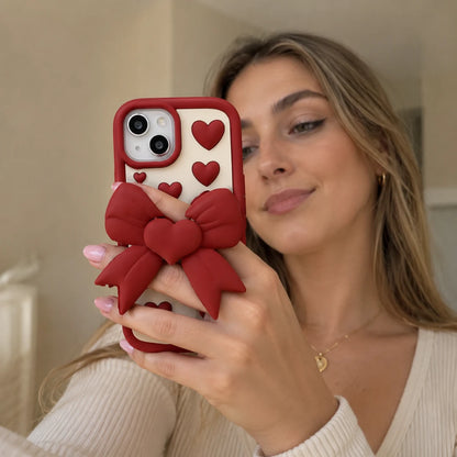 Josh & Tate - LoveBow™ Anti-Drop Phone Case - A woman holding a red phone case with a large red bow and heart details, taking a mirror selfie in an indoor setting.