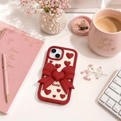 Josh & Tate - LoveBow™ Anti-Drop Phone Case - A red and cream phone case with a 3D red bow and heart pattern, displayed on a styled desk next to a pink notebook, a keyboard, and a pink coffee mug.