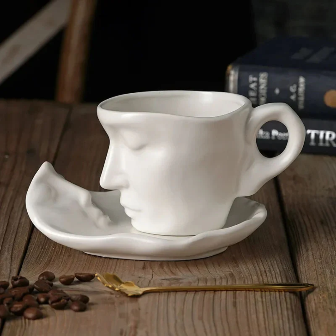White ceramic cup shaped like a face on a wooden table with coffee beans and a spoon.