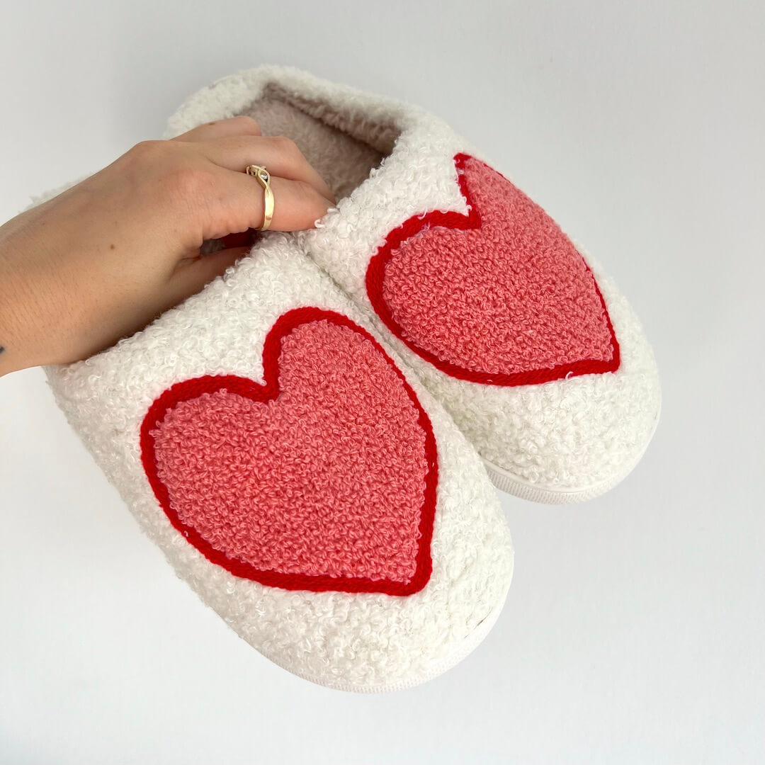 White slippers with red heart designs held by a hand on a light gray background