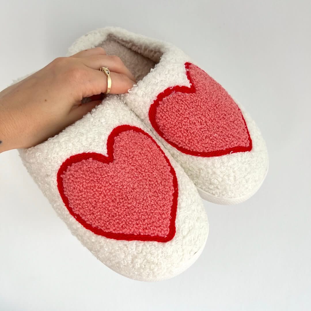 White slippers with red heart designs held by a hand on a light gray background
