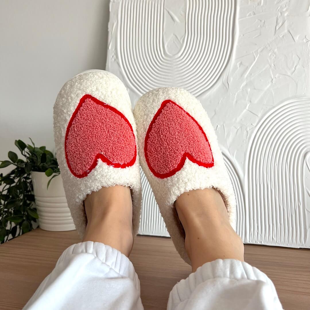 White slippers with red heart designs worn indoors against a textured white wall.
